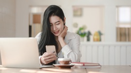  Een foto van een buitenlandse vrouw die glimlacht terwijl ze naar haar telefoon kijkt, met een laptop en een kop koffie voor haar