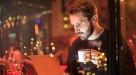 Een foto van een man in een café die een kopje koffie vasthoudt terwijl hij naar iets op zijn laptop kijkt